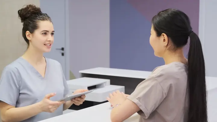 A clinic receptionist holding a tablet and talking with a visitor at the front desk