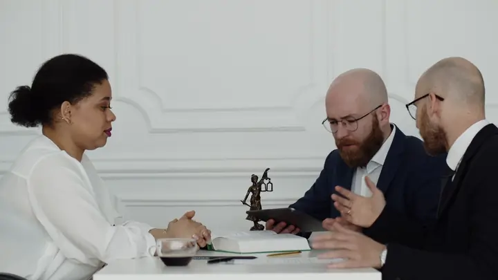 Three people in a bright office meeting around a white table with a small statue of Lady Justice