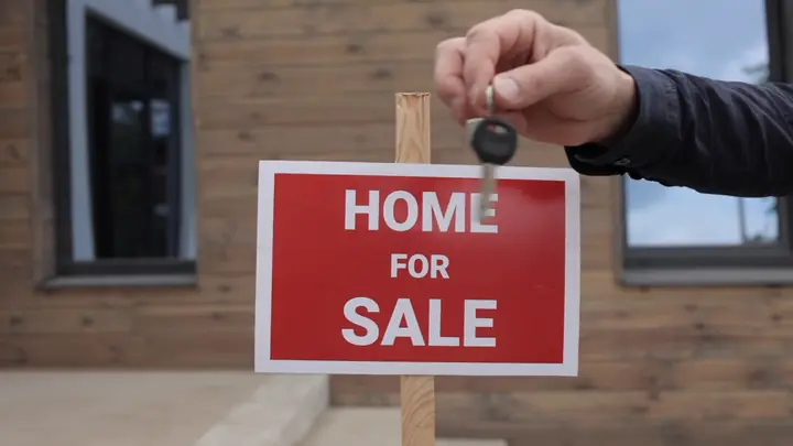 A hand holding out a house key in front of a Home for Sale sign